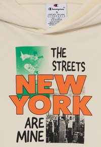 Krém színű kapucnis pulóver, merész fekete és narancssárga grafikákkal. A szöveg ezt olvassa: "NEW YORK UTCÁI AZ ENYÉMek" egy zöld és monokróm háttér előtt.