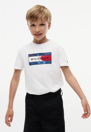 Blond jongetje draagt een wit T-shirt met een blauw-wit-rood rechthoekig logo en donkere korte broek tegen een effen witte achtergrond.