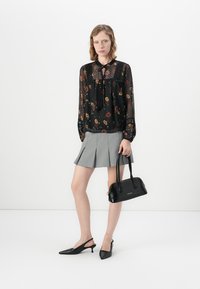 Donna in una blusa nera trasparente a fiori, gonna mini plissettata grigia, tacchi slingback neri, che tiene in mano una borsa nera su sfondo bianco.