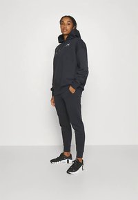 Sweatshirt preta com cordão ajustável, mangas compridas e bolso frontal, combinada com calças de treino a condizer e sapatilhas pretas.