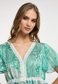 Abito verde trasparente con motivo paisley, scollatura a V, bordo in pizzo, maniche corte e vita con cordino. Tessuto leggero con dettagli floreali.