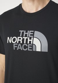 Schwarzes T-Shirt mit einem großen grauen und weißen "The North Face"-Logo, das auf der Brust aufgedruckt ist, getragen von einer Person mit heller Haut.