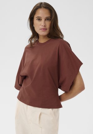 Femme aux cheveux bruns portant un haut ample à manches courtes marron et un pantalon beige clair, debout avec une main sur la hanche.