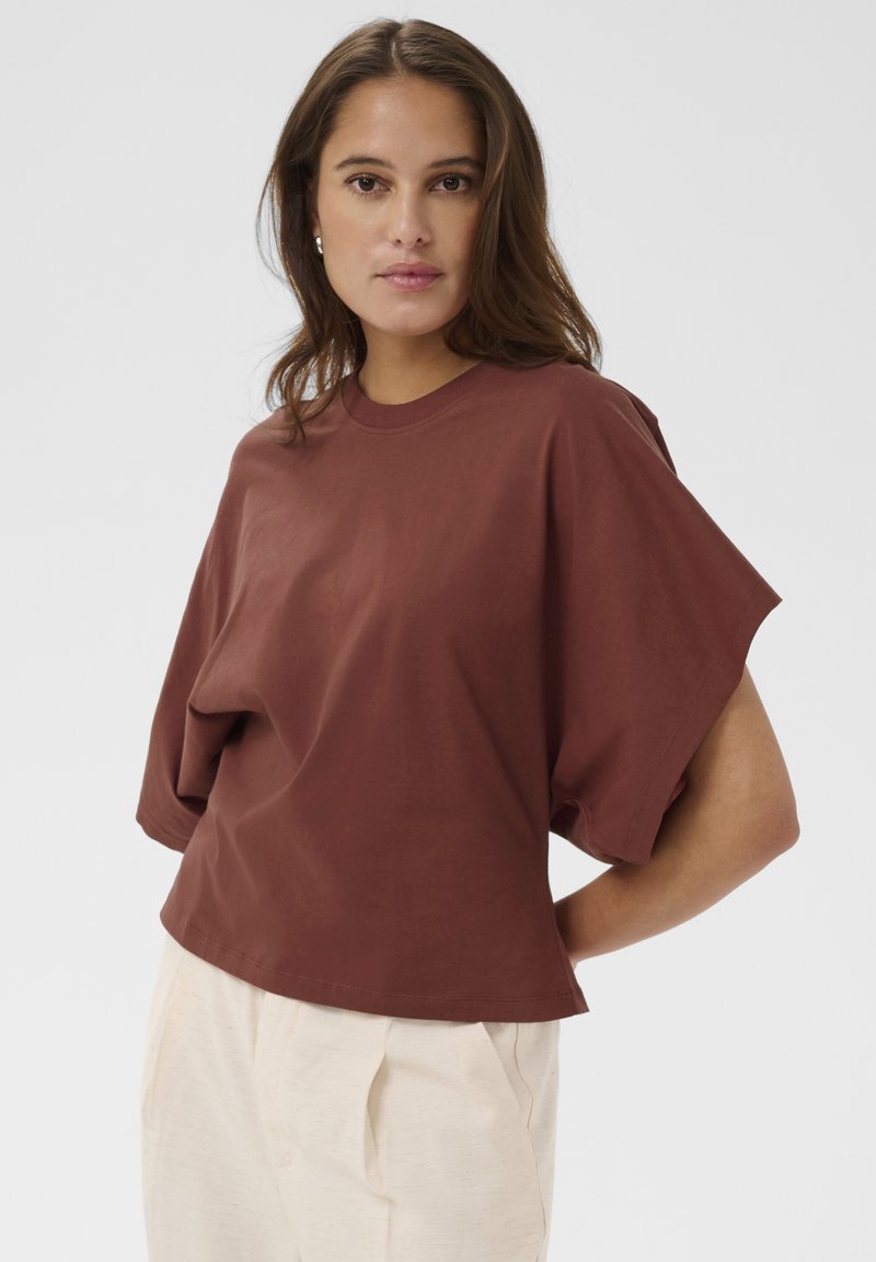 Femme aux cheveux bruns portant un haut ample à manches courtes marron et un pantalon beige clair, debout avec une main sur la hanche.