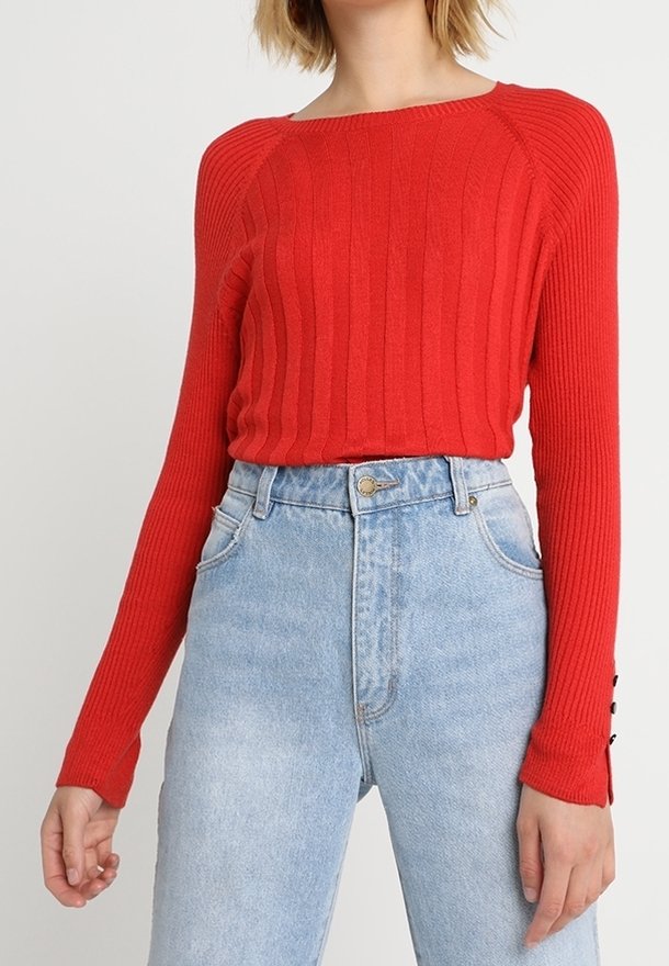 Femme portant un pull à manches longues côtelé rouge et ajusté, rentré dans un jean taille haute bleu clair avec fermeture par bouton et zip.