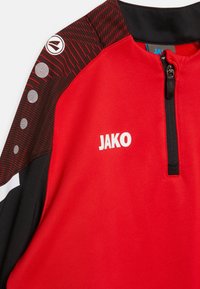 Rode sporttop met een zwarte kraag, voorzien van een kwart ritssluiting, gestreept patroon op de schouder en een wit logo "JAKO".