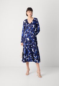 Μακρυμάνικο ναυτικό φόρεμα με αφηρημένο μπλε και λευκό μοτίβο, με ντεκολτέ σε V, φουσκωτή φούστα, φτιαγμένο από απαλό ύφασμα, συνδυασμένο με γυμνές ψηλοτάκουνες σαγιονάρες.