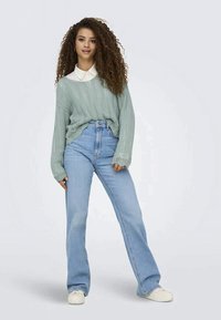 Young woman with long curly hair wears a light green knit sweater over a white collared shirt and light blue high-waisted jeans with white sneakers.