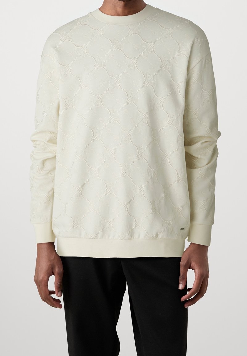 Homme portant un pull crème texturé à manches longues avec un col rond et un pantalon noir, sur un fond uni.