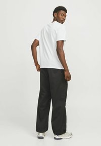 Witte katoenen t-shirt met een ronde hals, gecombineerd met losse zwarte broeken. Het model staat met de rug naar de camera, waardoor het ontwerp aan de achterkant zichtbaar is.