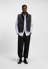 Gilet imbottito nero sopra una camicia a righe bianche e blu, abbinato a pantaloni larghi neri e scarpe stringate nere. Design semplice e moderno.