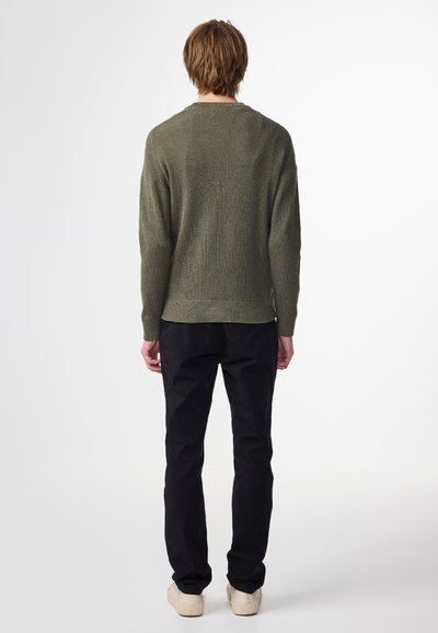 Maglione lavorato a maglia verde oliva con una texture a coste, collo rotondo e polsini aderenti, abbinato a pantaloni neri e scarpe beige.