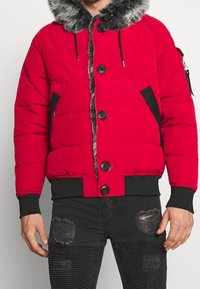 Rød puffjakke med pelsforet hætte, sorte detaljer, frontknapper og sidelommer, parret med slidte sorte jeans.