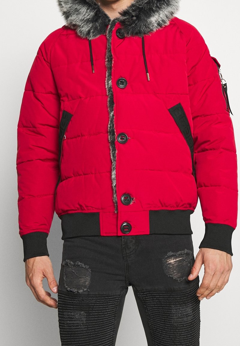 Rød puffjakke med pelsforet hætte, sorte detaljer, frontknapper og sidelommer, parret med slidte sorte jeans.