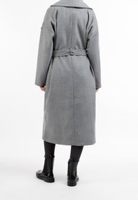 Manteau long en laine grise avec une ceinture à la taille, de larges revers et des manches longues, associé à des bottines noires et des leggings foncés.