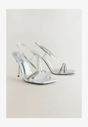 Srebrne sandale na visokom petu z adjustable trakami, z kvadratnimi prsti in okrasnimi kamenčki na sprednji strani. Gladek metalni zaključek.