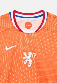 Oransje Nike-trøye med et hvitt løveemblemet, teksturert prikkemønster og en flerfarget ribbestrikket krage med røde, hvite og blå aksenter.