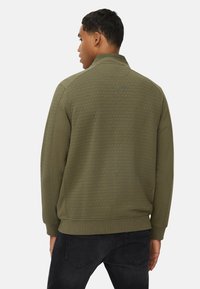 Olijfgroene sweatshirt met een gestructureerd hexagonaal patroon, voorzien van een kraag en geribde manchetten, gedragen over donkere jeans.