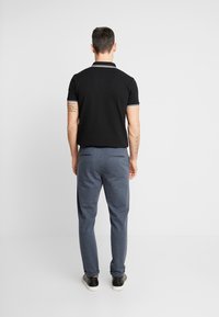 Mies, der ein schwarzes Polohemd und eine blaue Hose trägt, steht mit entspannten Armen an den Seiten einem schlichten weißen Wand gegenüber.