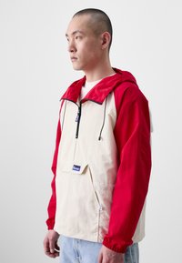 Roter und beiger Pulloverjacke mit Kapuze, Fronttasche, Reißverschlussakzenten, elastischen Bündchen und einem Etikett auf der Tasche. Glatte Textur.