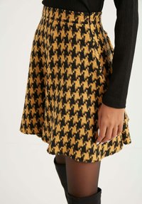 Mujer que lleva una falda de estampado de pied de poule en amarillo y negro, una blusa negra de mangas largas, medias transparentes y botas altas negras hasta la rodilla.