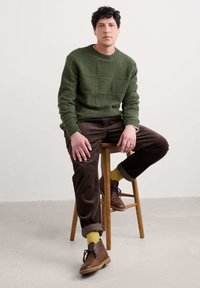 Maglione verde scuro con motivo quadrato testurizzato, pantaloni in velluto a coste marroni, calze gialle e scarpe marroni, seduto su uno sgabello di legno.