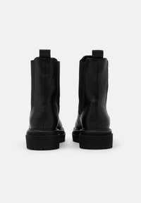 Bottines en cuir noir avec semelles épaisses et languettes, montrées de dos sur un fond blanc uni.