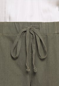 Gros plan sur un pantalon vert olive avec un cordon en tissu noué à la taille, porté sur une chemise blanche.