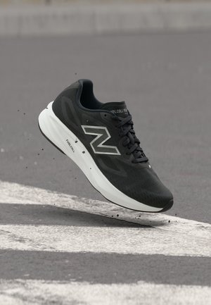 Chaussure de running New Balance noire avec semelle blanche FuelCell, capturée en plein air au-dessus d'une surface en asphalte gris avec des marquages routiers blancs.