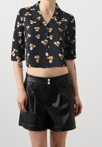 Chemisier noir à fleurs, court, avec des manches trois-quarts, orné de fleurs blanches et orange, associé à un short en cuir noir plissé taille haute.