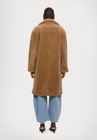 Bruine, fuzzy wollen jas met een brede kraag en een rechte snit, gecombineerd met lichtblauwe wijde jeans en zwarte sandalen met bandjes.