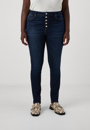 Dark blue, high-waisted jeans with a button-front closure, five pockets, and a slim fit. Worn with patterned footwear.