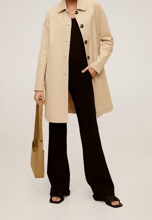 Personne portant un manteau beige boutonné par-dessus une tenue noire, tenant un sac à main beige, et portant des sandales noires ouvertes aux orteils, debout devant un fond neutre.