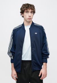 Ναυτικό μπλε αθλητικό μπουφάν από λείο ύφασμα, με λευκές ρίγες στους ώμους και μικρό λογότυπο στο αριστερό στήθος. Φερμουάρ μπροστά.