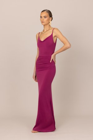 Kvinne iført en tettsittende, ermeløs gulvlengde kjole i magenta med tynne stropper, gullkjede og matchende blomsterøreringer, stående på siden.