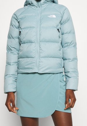 Doudoune courte bleu clair avec fermeture éclair et logo sur la poitrine, portée avec une jupe assortie bleu clair à ourlet arrondi.