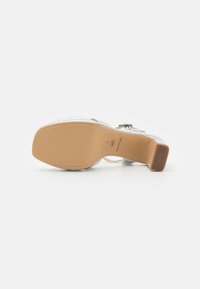 Sandalia de tacón alto plateada con un upper suave, correas transparentes y una suela contorneada beige que presenta un agarre texturizado y un logo.
