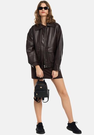 Kvinde iført mørkebrun læderjakke, sorte solbriller, sort kjole og sorte sneakers, der holder en lille sort rygsæk i stropperne.