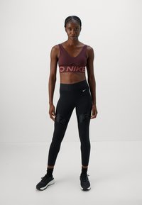 Sportos nő áll szemben, bordó Nike sportmelltartót és fekete Nike Pro leggingset visel, fekete sportcipővel.