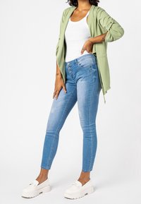 Jasnozielony otwarty kardigan na białym topie na ramiączkach, w połączeniu z niebieskimi skinny jeans z zapięciem na guziki oraz białymi platformowymi loafersami.