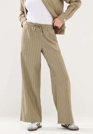 Osoba nosząca beżowe spodnie w szerokie prążki z elastycznym pasem ze sznurkiem, białe trampki z czarnymi paskami oraz pasującą kurtkę.