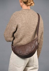 Bolso de hombro de cuero marrón con forma curva y textura suave, llevado de lado, combinado con un suéter de punto beige y pantalones claros.