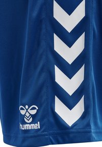 Blått stoff med hvite nedoverpekende chevronstriper og en hvit hummel-logo nær kanten på et sportplagg.