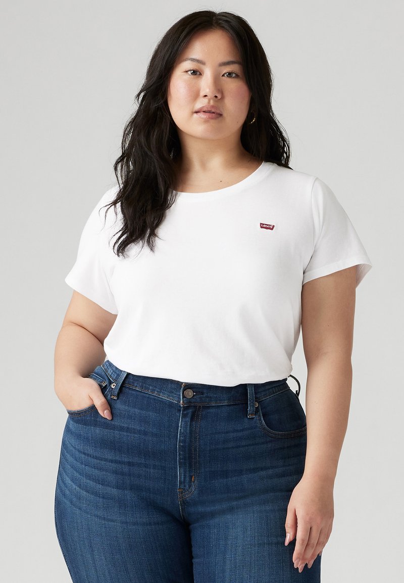 Camiseta de algodón blanca con escote redondo y mangas cortas, que presenta un pequeño logo rojo de "Levi's" en el lado izquierdo del pecho, combinada con jeans de color azul oscuro.