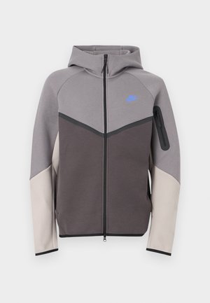 Gray and beige zip-up hoodie with a hood. Features a blue logo, contrast seams, and a zippered pocket on the sleeve. Smooth texture.