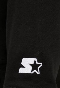 Černá látka s bílou vyšívanou logem se "S" a hvězdičkou. Textura je hladká s mírným elastickým efektem. Detailní záběr.
