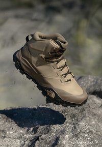 Beżowe buty trekkingowe z wsparciem kostki, strukturalna cholewka, wzmocniony czubek i czarna gumowa podeszwa. Posiada brązowe sznurówki i detal z logo na boku.