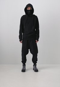 Schwarzer Hoodie mit Kordelzug, kombiniert mit lockeren schwarzen Hosen mit Taschen. Getragen mit gemusterten schwarzen Stiefeln. Einfaches, modernes Design.