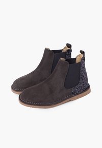 Botas Chelsea de ante marrón oscuro con paneles de talón negros brillantes, inserciones elásticas en los lados y suelas de goma marrón claro, mostradas en un par lado a lado.