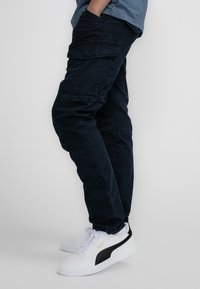 Donkerblauwe cargobroek met meerdere zakken, gemaakt van stevig materiaal, gecombineerd met witte sneakers met zwarte en gouden accenten.